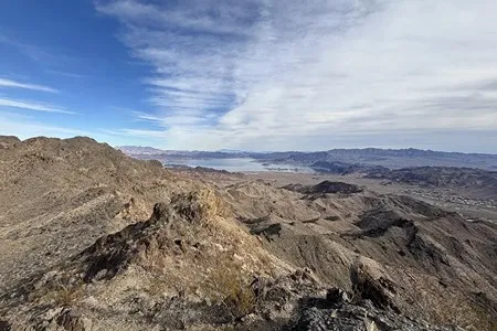 A panoramic view from a rocky mountain ridge overlooking a vast, dry desert landscape with distant blue water and hazy mountains under a partly cloudy sky.