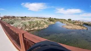 A view from a bridge looking out over a slow-moving river with lush green and silver vegetation lining its banks under a blue sky with scattered clouds.
