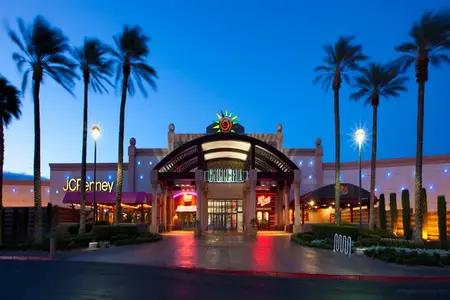 The brightly lit entrance of a large shopping mall at dusk, featuring palm trees lining the walkway and various store signs illuminated against the dark blue sky.