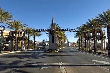An archway over a street reads "HISTORIC DOWNTOWN" in large metal letters, flanked by palm trees and leading into a sunny downtown area with shops and vehicles under a clear blue sky.