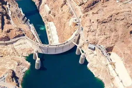 An aerial view of the Hoover Dam, a massive concrete arch-gravity dam spanning a deep blue river gorge in a dry, rocky landscape under a bright sky.