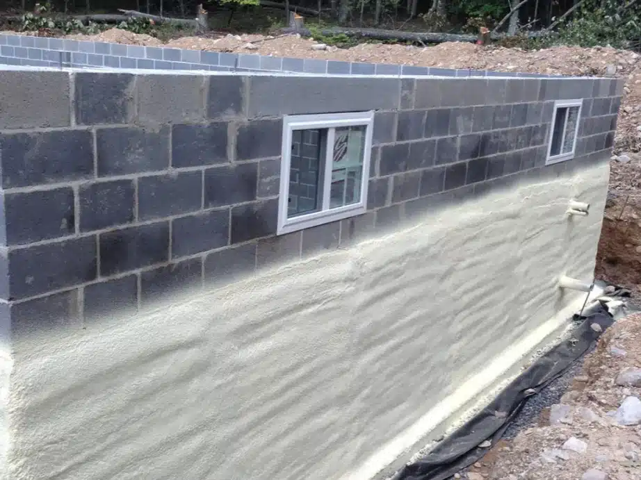 The exterior of a concrete block foundation wall shows a layer of light beige spray foam insulation applied to its surface. Two small windows with white frames are visible in the block wall above the insulation.