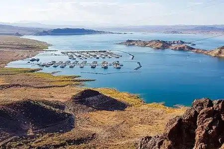 A panoramic view of a vast blue lake dotted with islands and surrounded by arid, brown and tan hills under a hazy sky. A marina with many houseboats is visible along one shore.