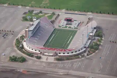 An aerial view of a large, open-air football stadium with green artificial turf, surrounded by empty parking lots and some greenery.